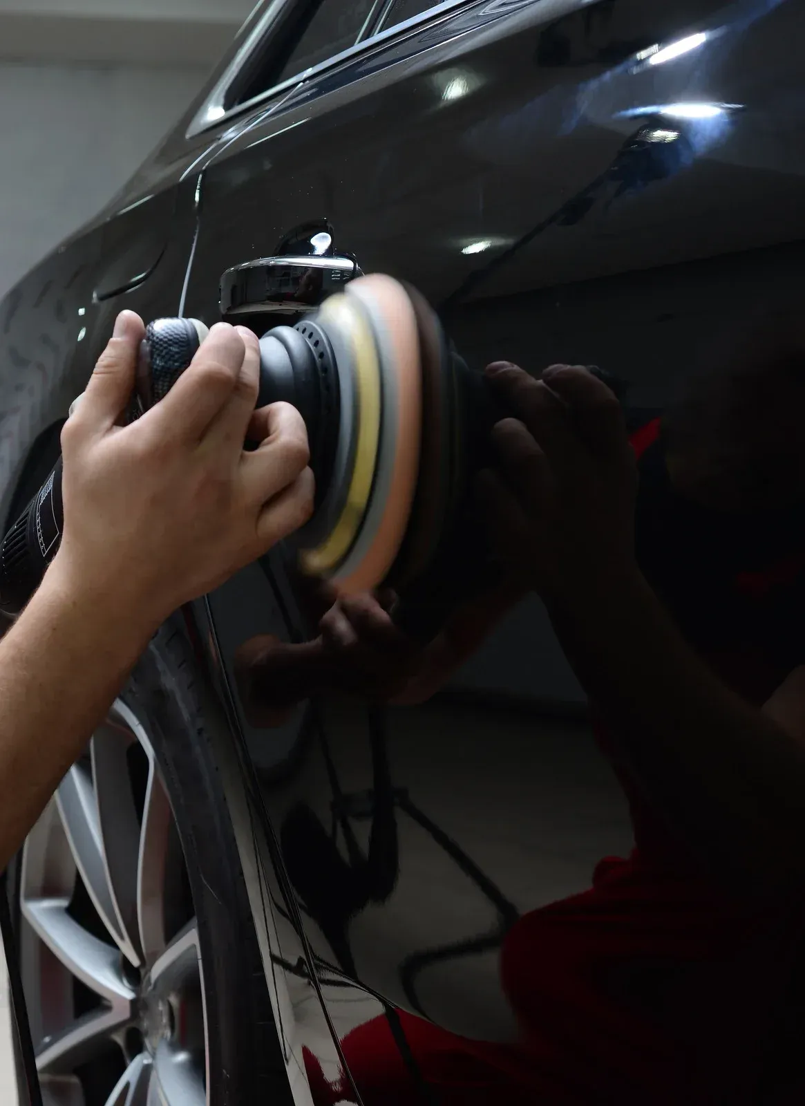 Person polishing a black car with an electric buffer in a garage.