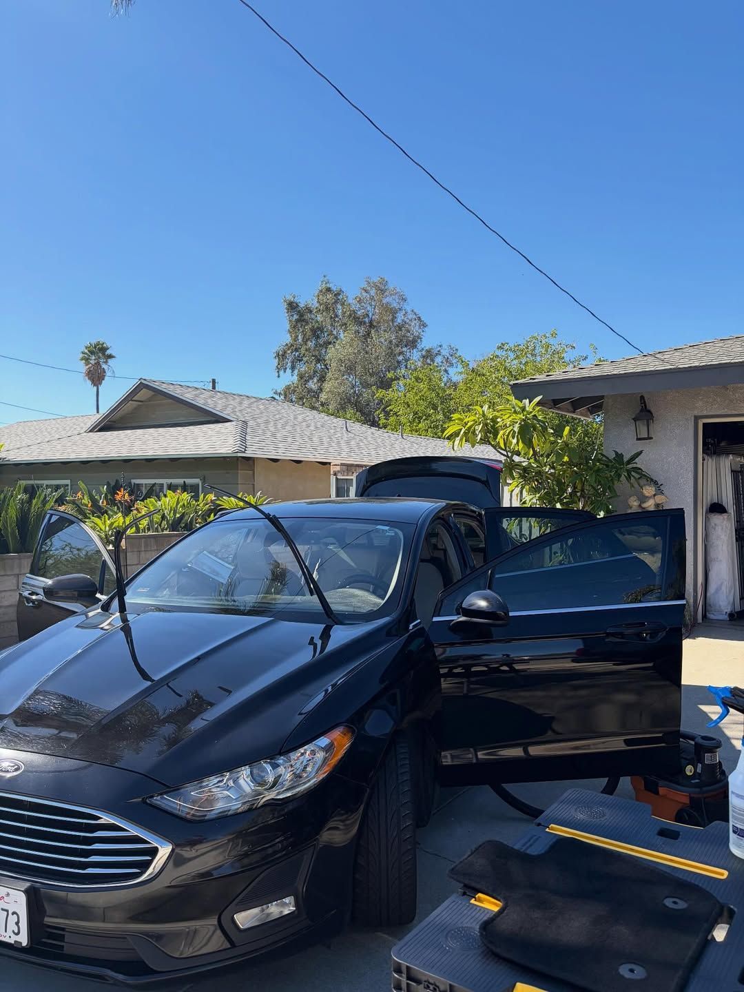 Black car with open doors, being cleaned outdoors on a sunny day.