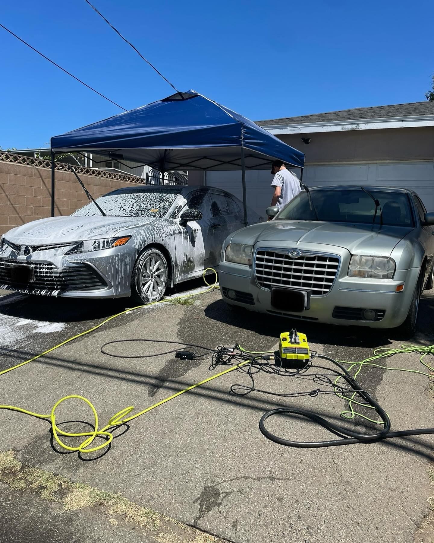 Two cars, a silver Camry and a Chrysler 300, being washed under a blue canopy. Person washing the Camry.