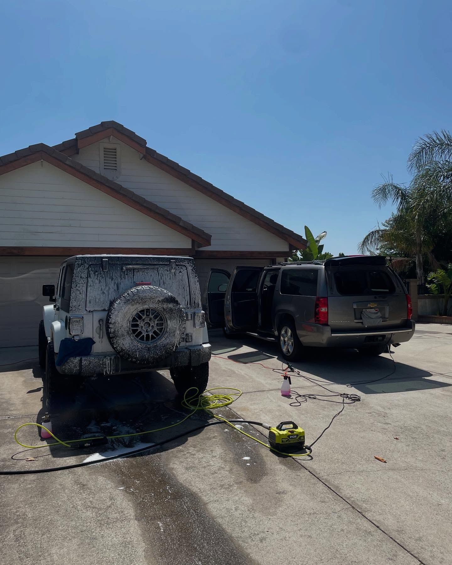 Jeep and SUV being washed in a driveway on a sunny day. Soap covers the Jeep; the SUV's doors are open.