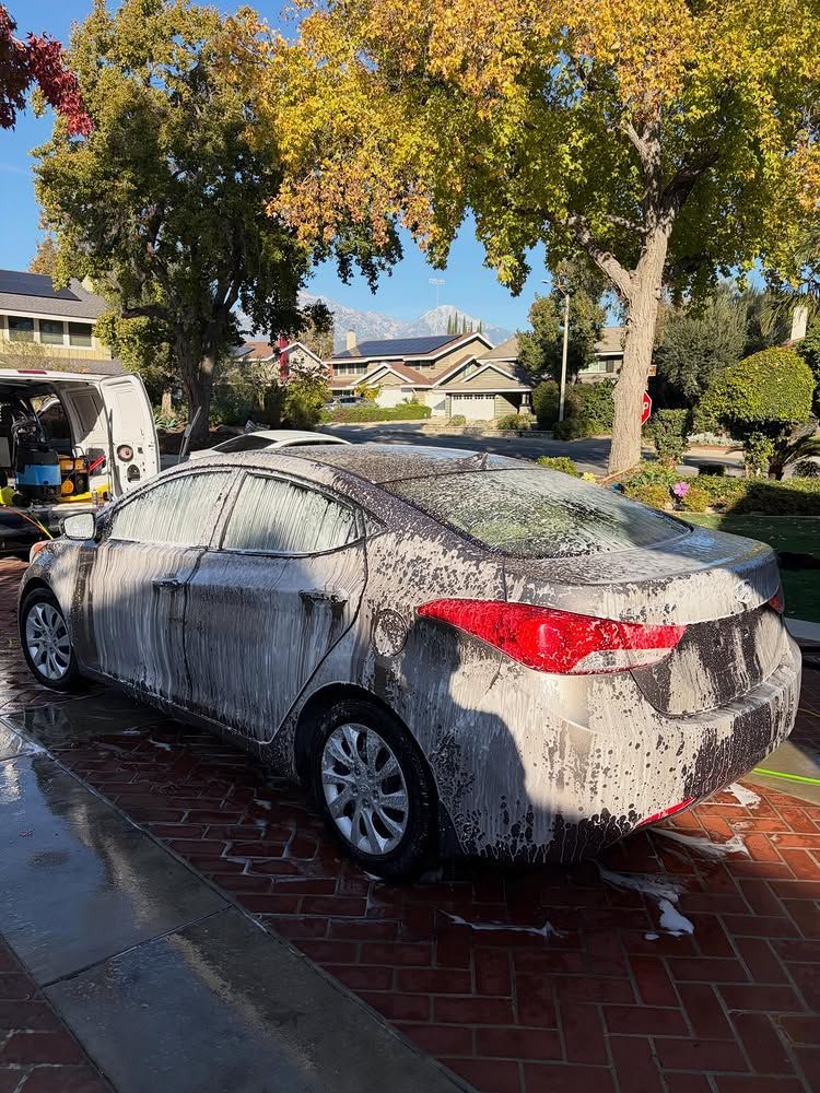 Gray sedan covered in white foam being washed on a brick driveway, with trees and houses in the background.