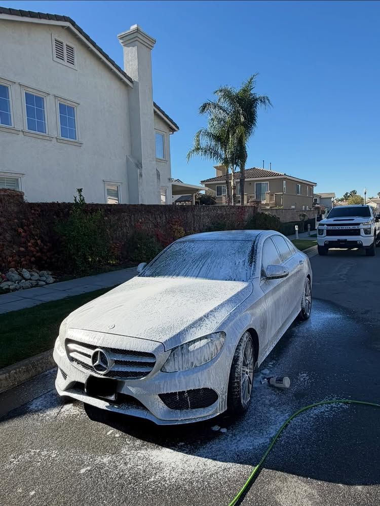 White Mercedes car covered in foam being washed on a street in front of a house on a sunny day.