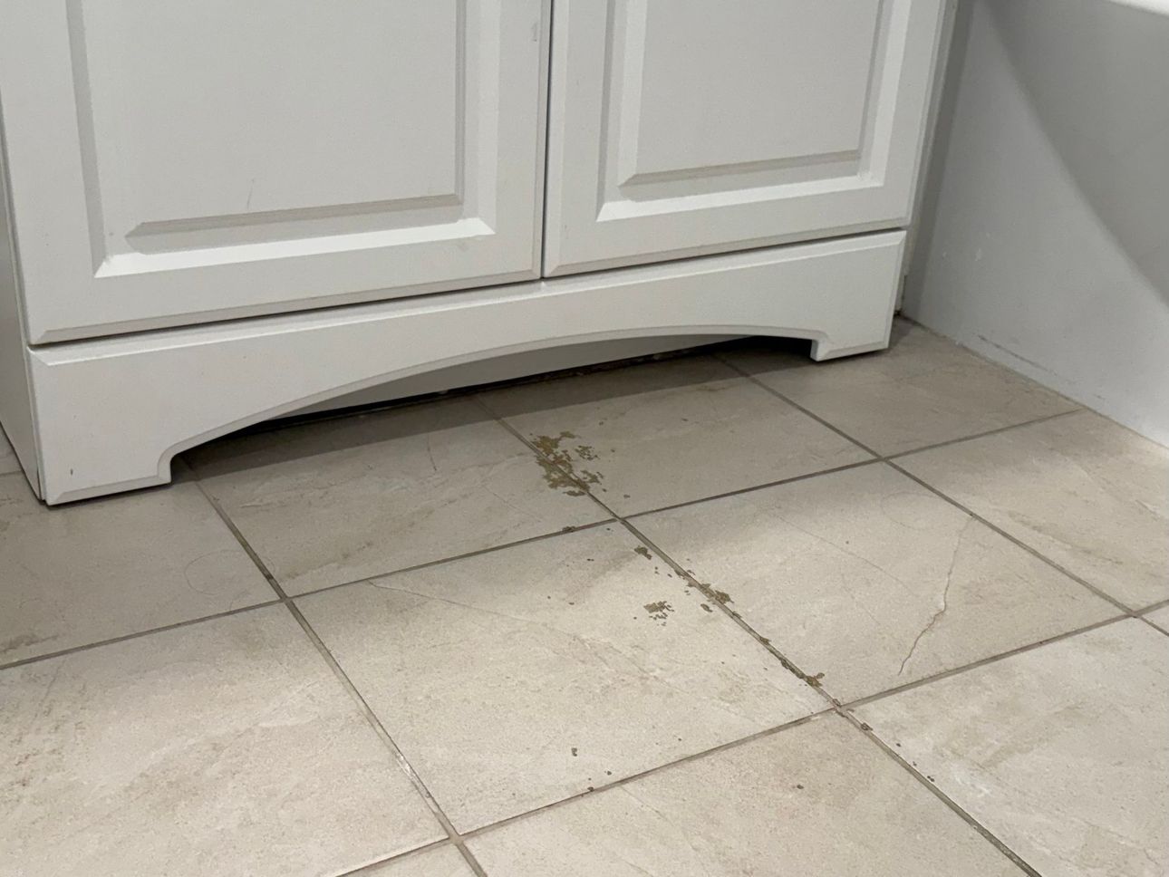 White bathroom cabinet base with water stain on tan tile floor.