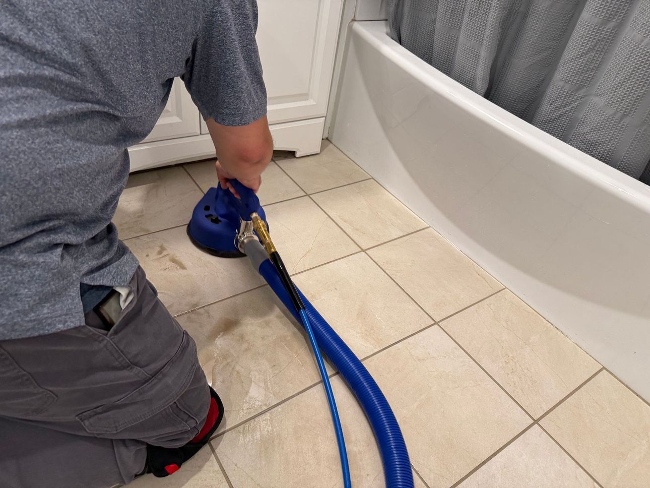Person kneeling, cleaning bathroom floor with a blue and black cleaning tool, next to a white tub.