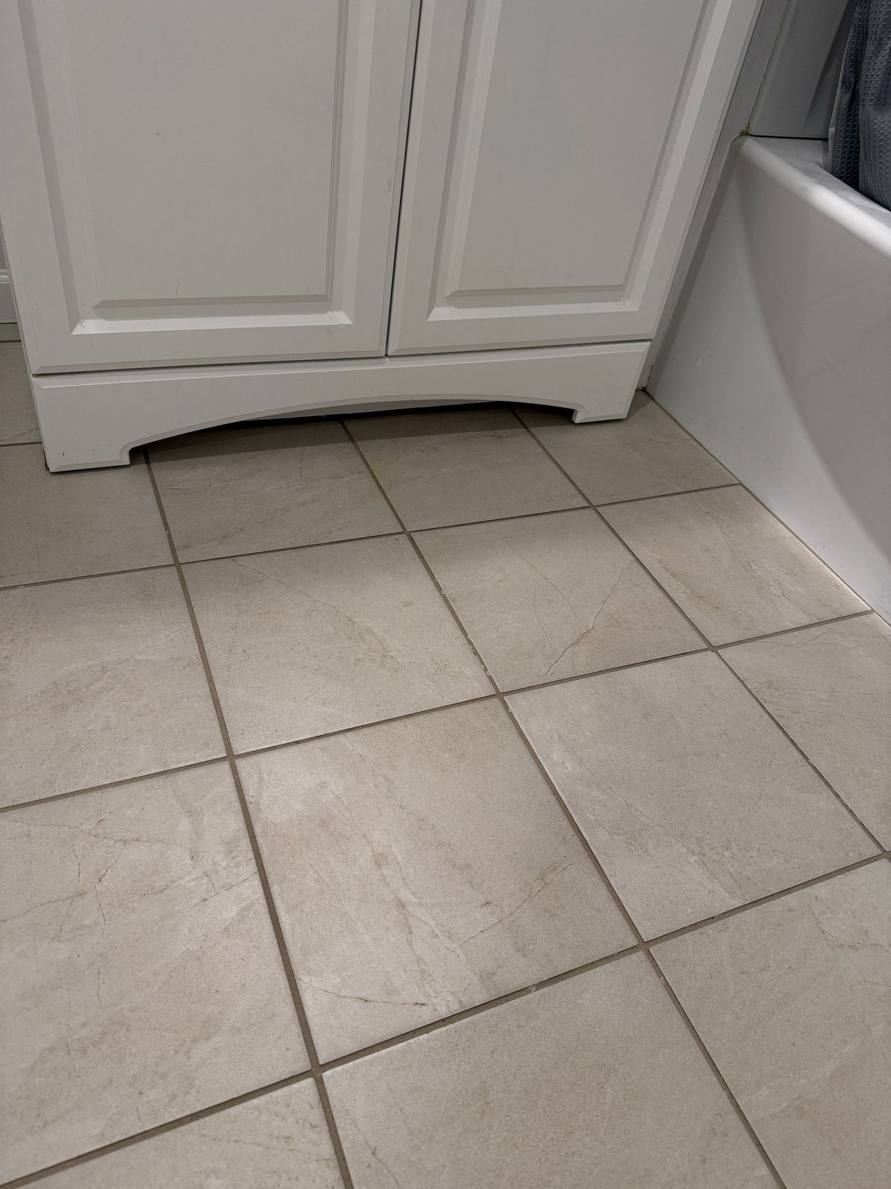 Beige tiled bathroom floor in front of a white cabinet, with the edge of a bathtub visible on the right.