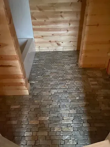 A bathroom with light wood-paneled walls and a rectangular white bathtub, featuring a rustic stone-tiled floor.