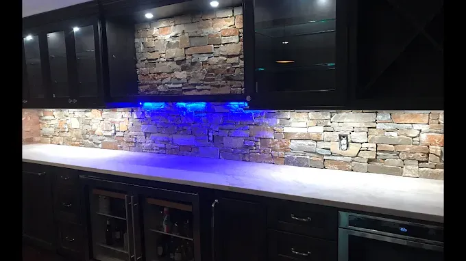 A modern wet bar featuring dark cabinetry, a light stone backsplash, a white countertop, and blue under-cabinet lighting.