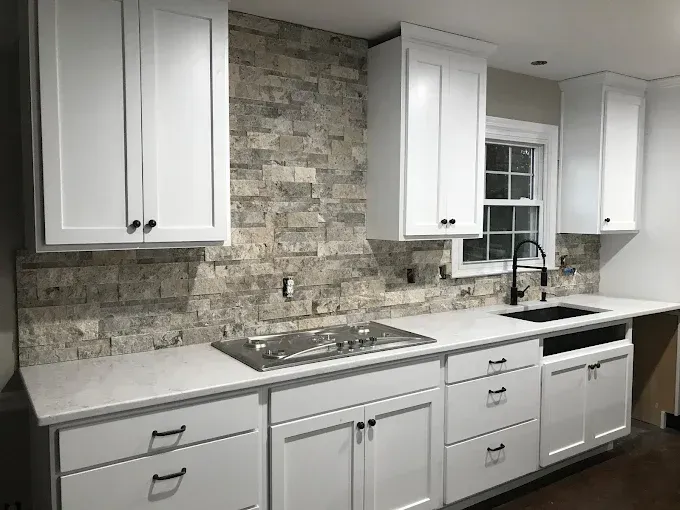 A modern kitchen with white cabinets, light-colored stone backsplash, white countertops, and a black faucet.