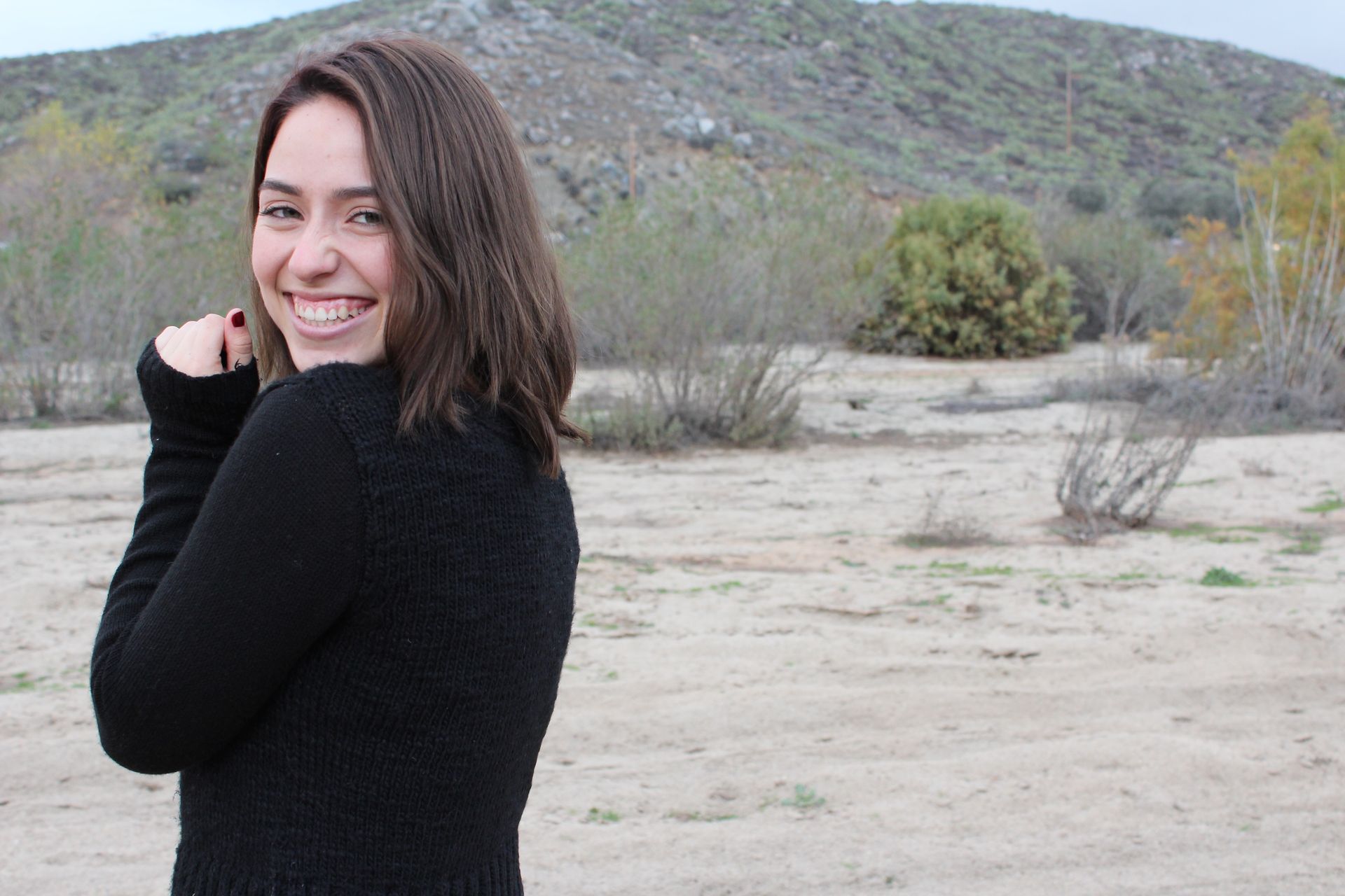Kayla in a black sweater smiles broadly outdoors, with brown fields and a hillside in the background.