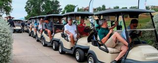 Golf carts lined up on a road, filled with people. A green golf course is visible in the background.