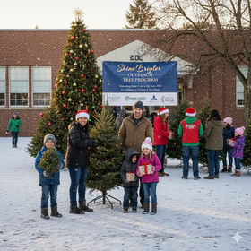People pose with Christmas trees in the snow outside a school building. A sign reads, 