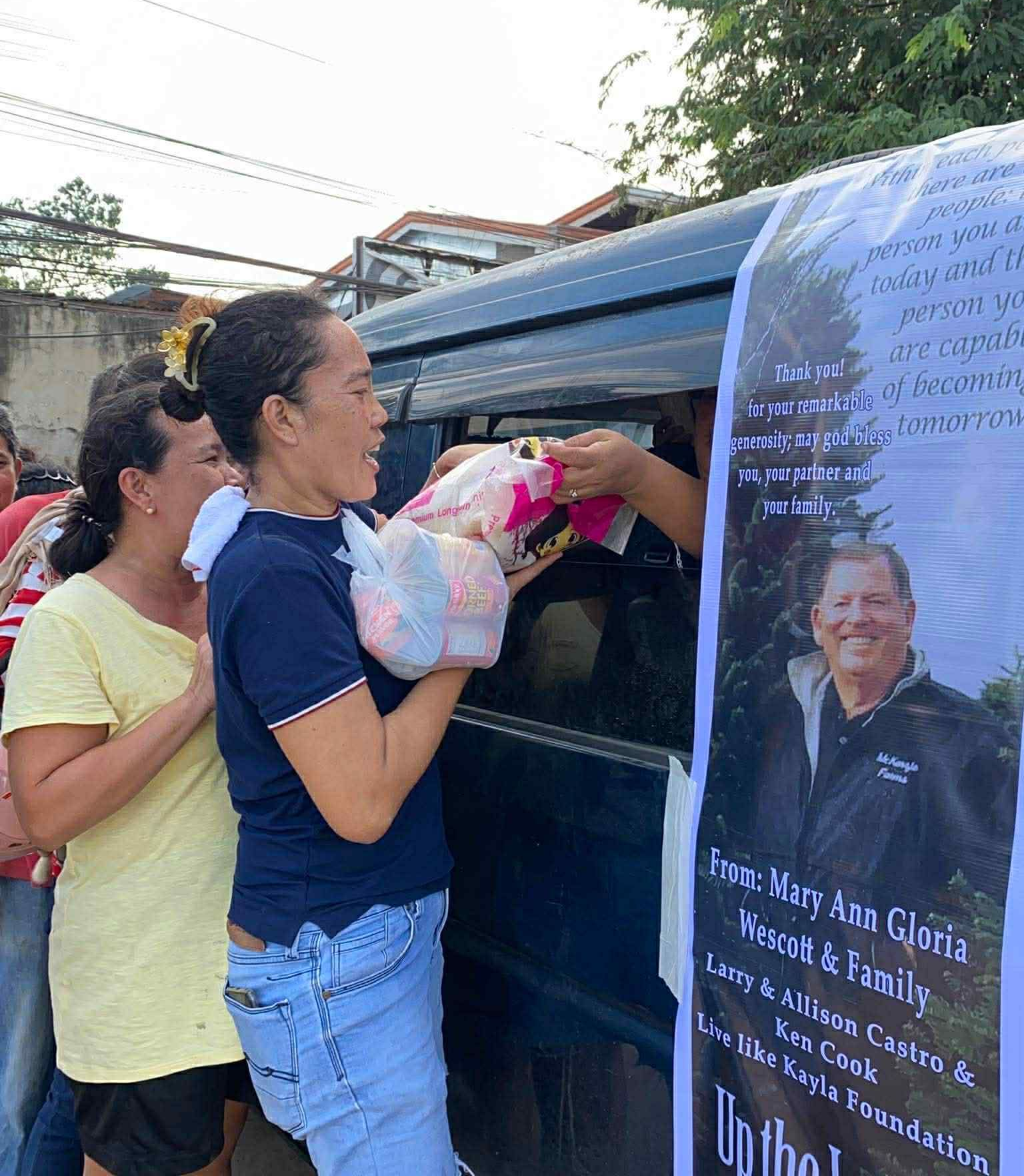People receiving donations from a vehicle. A woman hands a package to another woman. Sign with a photo of a man.