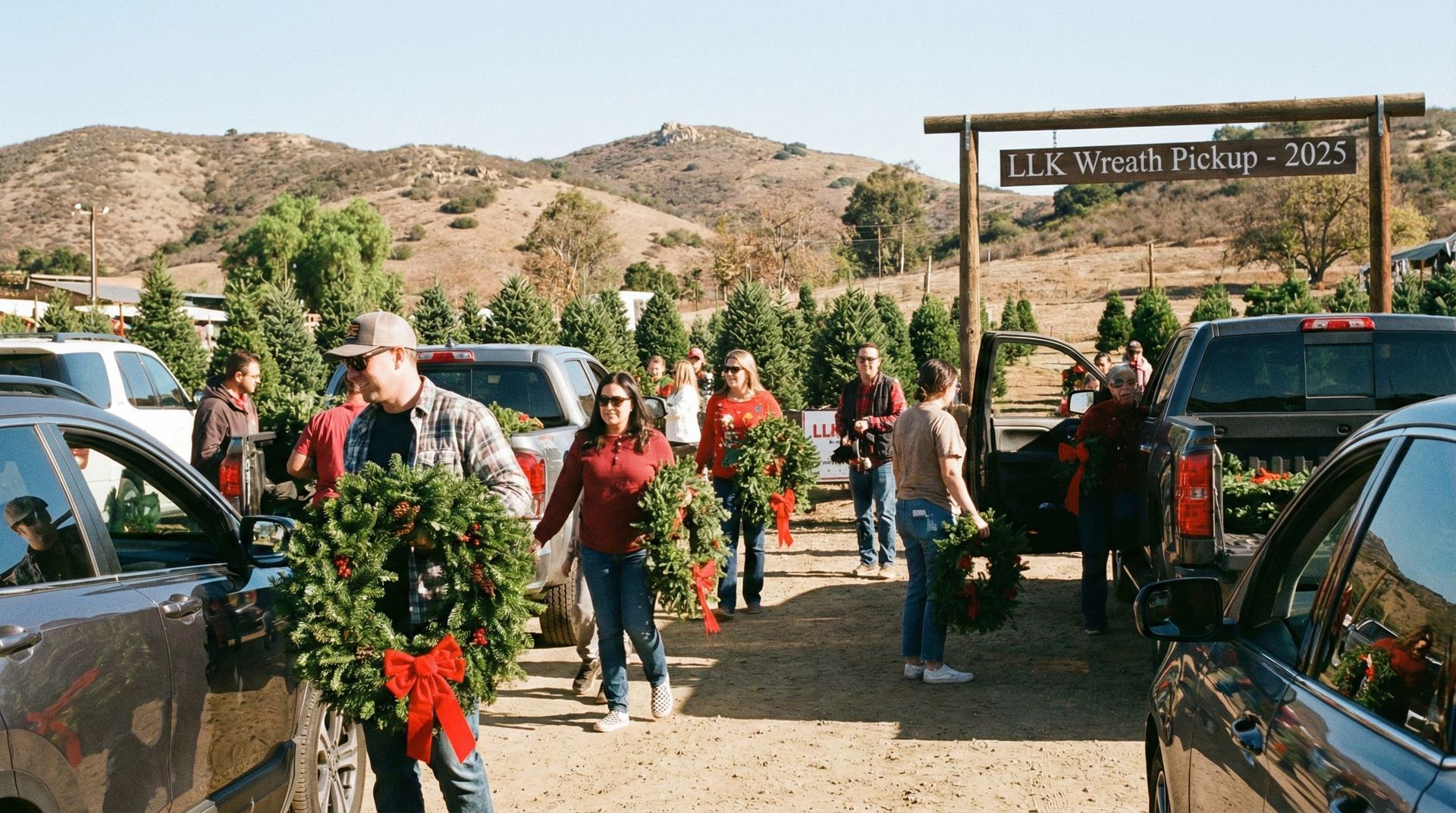People carrying wreaths at a Christmas tree farm entrance; vehicles parked. Sunny day, hills in background.
