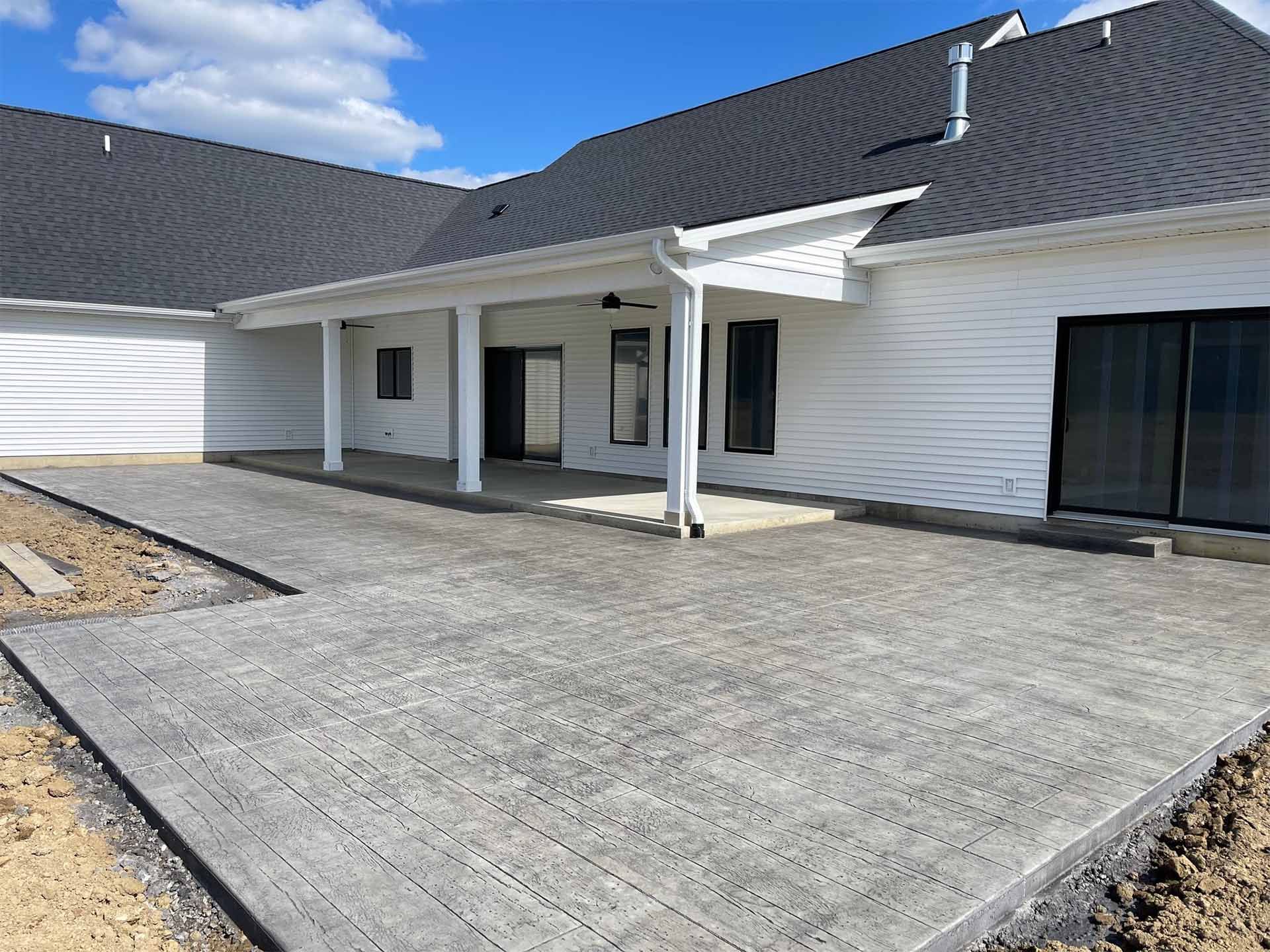 Newly poured concrete patio and walkway next to a white house with a dark roof and covered porch.