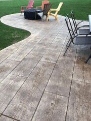 Wooden plank-textured concrete patio with outdoor furniture, fire pit, and a green lawn.