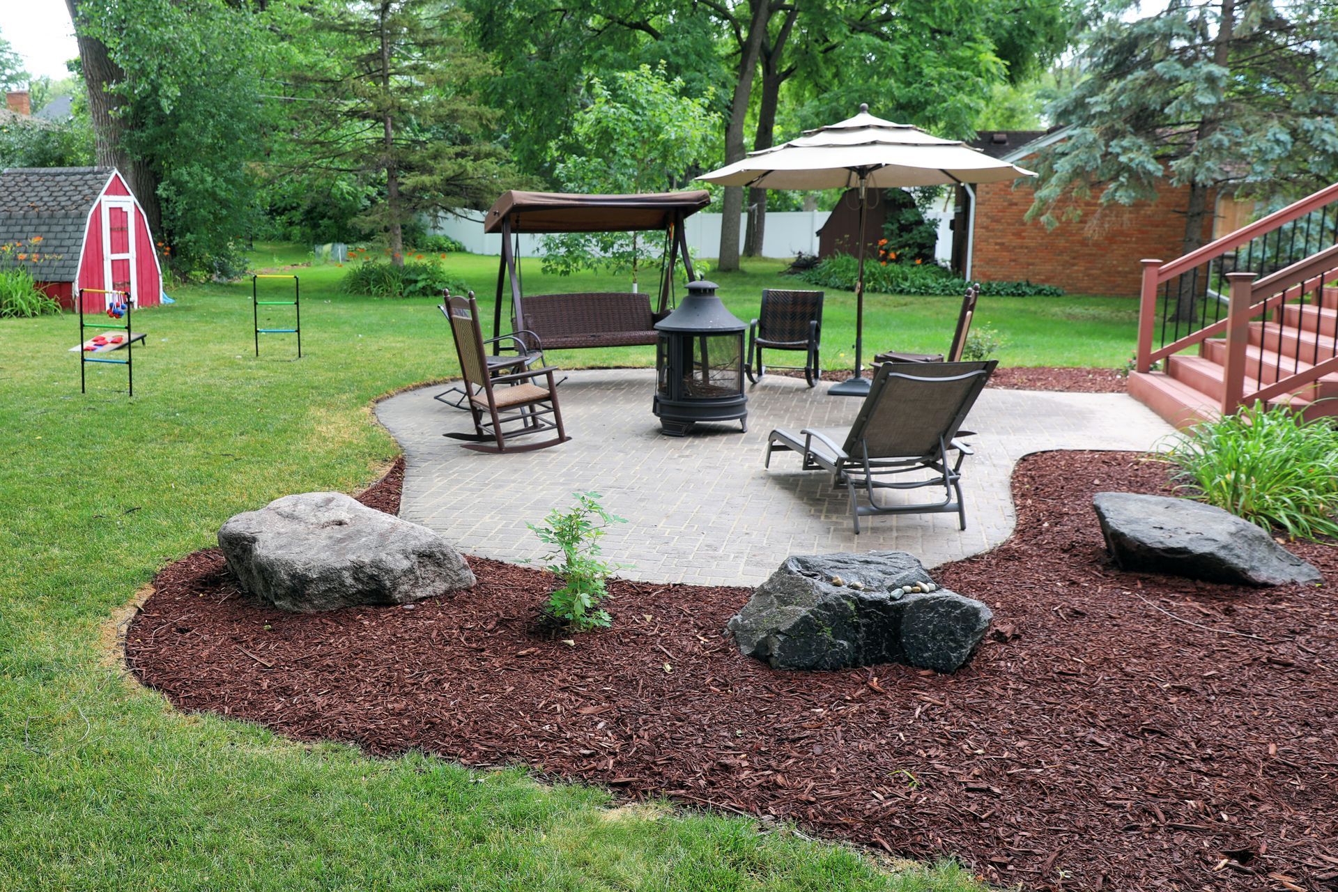 A backyard with a stamped patio, chairs, a swing, a fire pit, an open umbrella, and trees around.