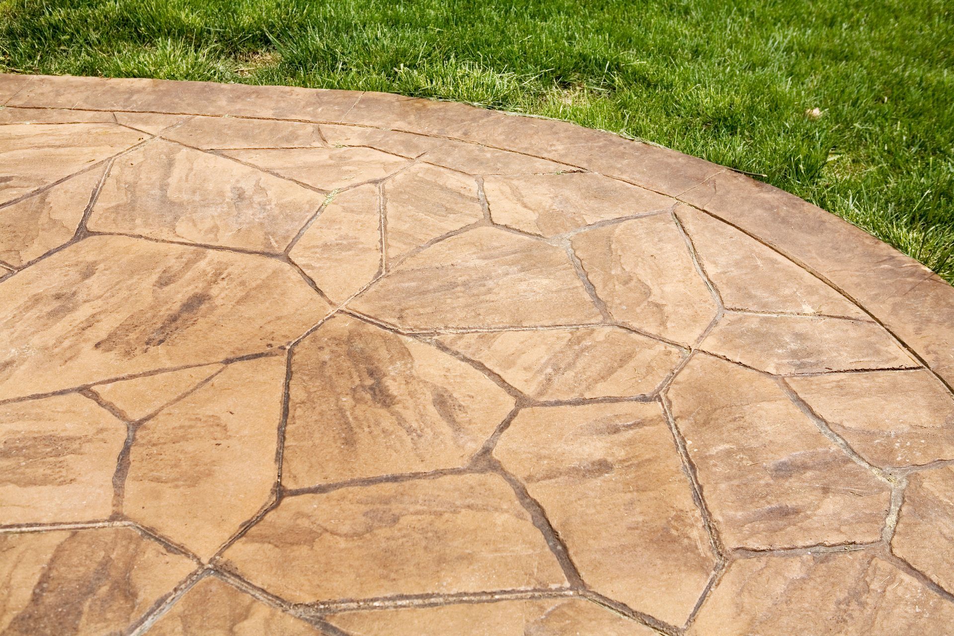 Stamped concrete patio with a stone-like pattern bordered by green grass in a backyard. Stamped concrete patio with a stone-like pattern bordered by green grass in a backyard.