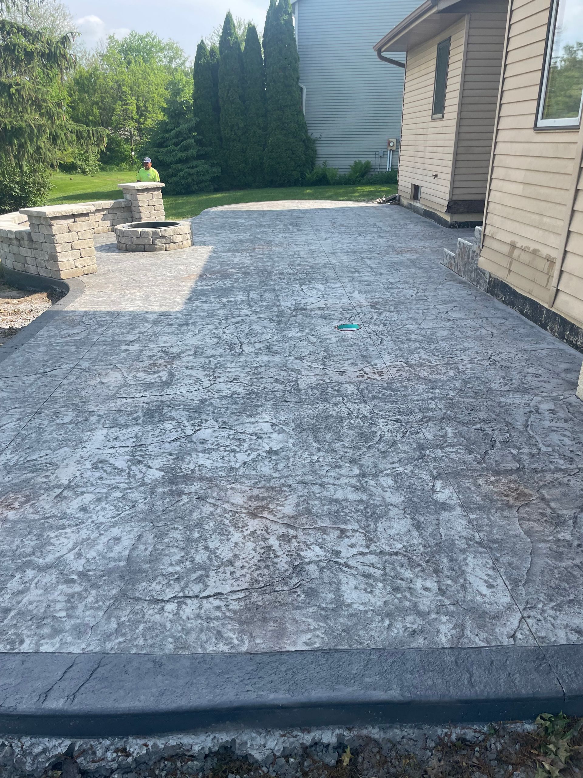 Concrete patio with fire pit, adjacent to a beige house. Gray stone border, green grass visible.