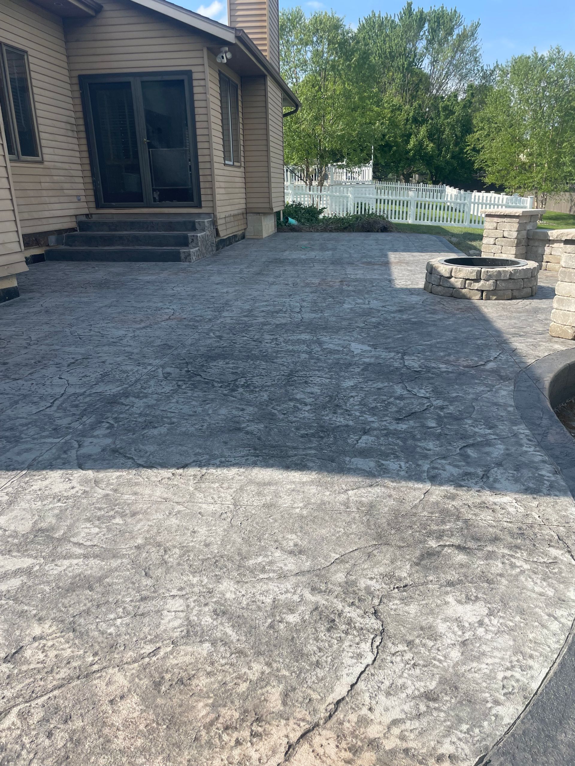 Concrete patio outside a house with dark gray textured finish, stone firepit, and steps leading to a glass door.