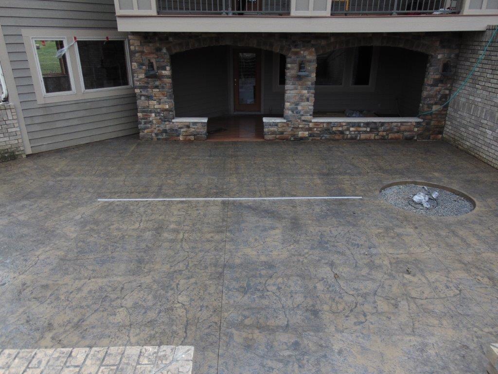 Stone-textured concrete patio in front of a house. Includes a fire pit, porch with stone pillars, and gray siding.