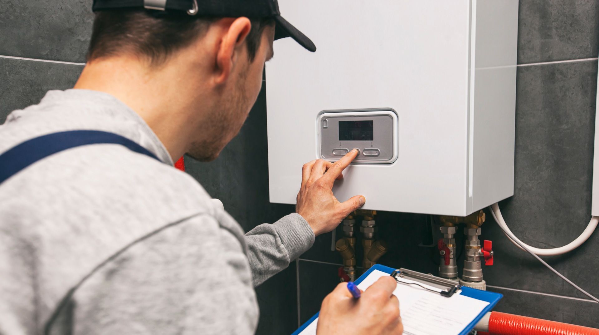 A person wearing a uniform cap and grey sweater adjusts a wall-mounted boiler control panel while holding a clipboard.