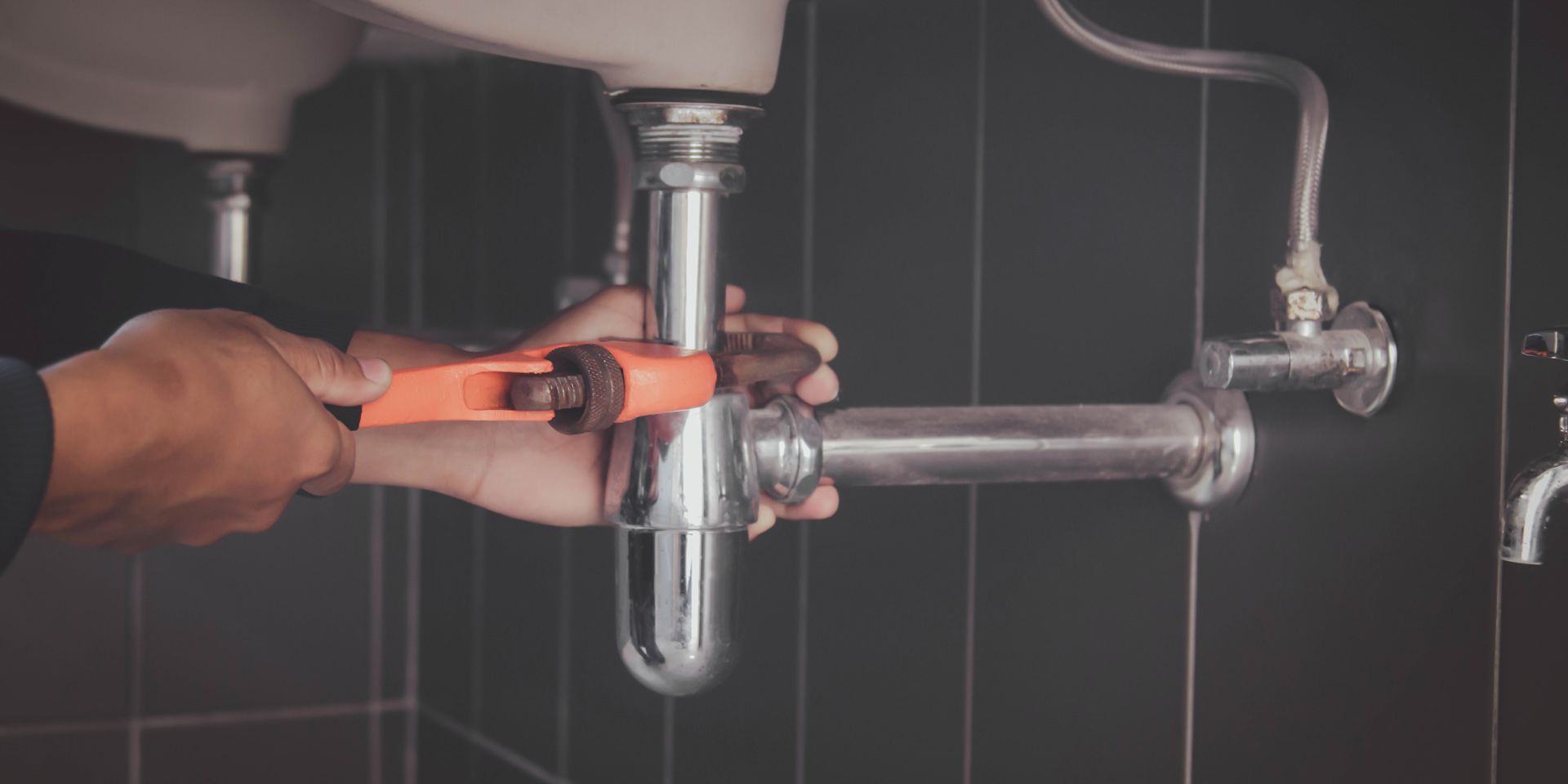 Hands use an orange pipe wrench to tighten a chrome p-trap under a bathroom sink against a dark tiled wall.