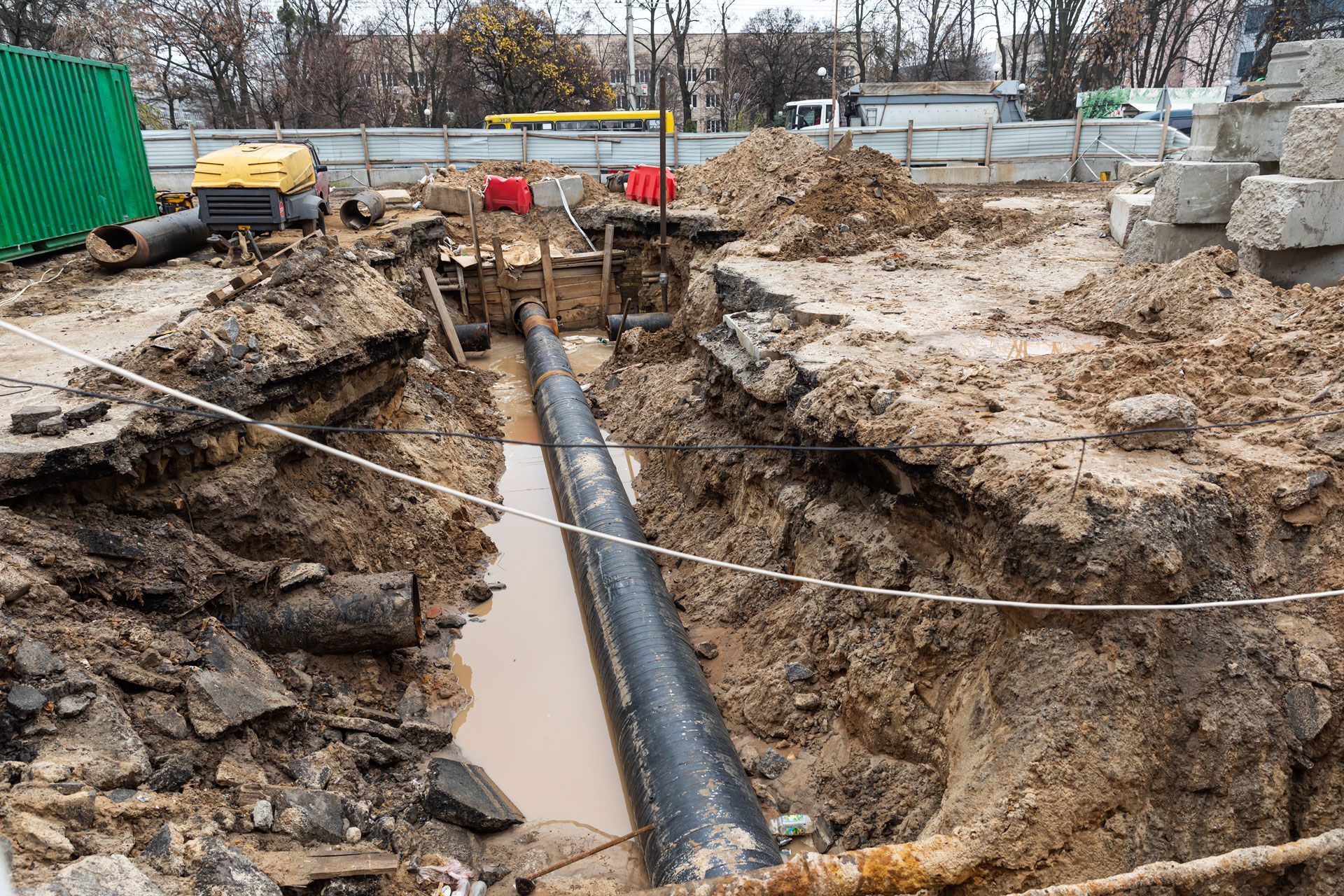 A deep construction trench with a long black pipe running through the center, surrounded by dirt and safety fencing.