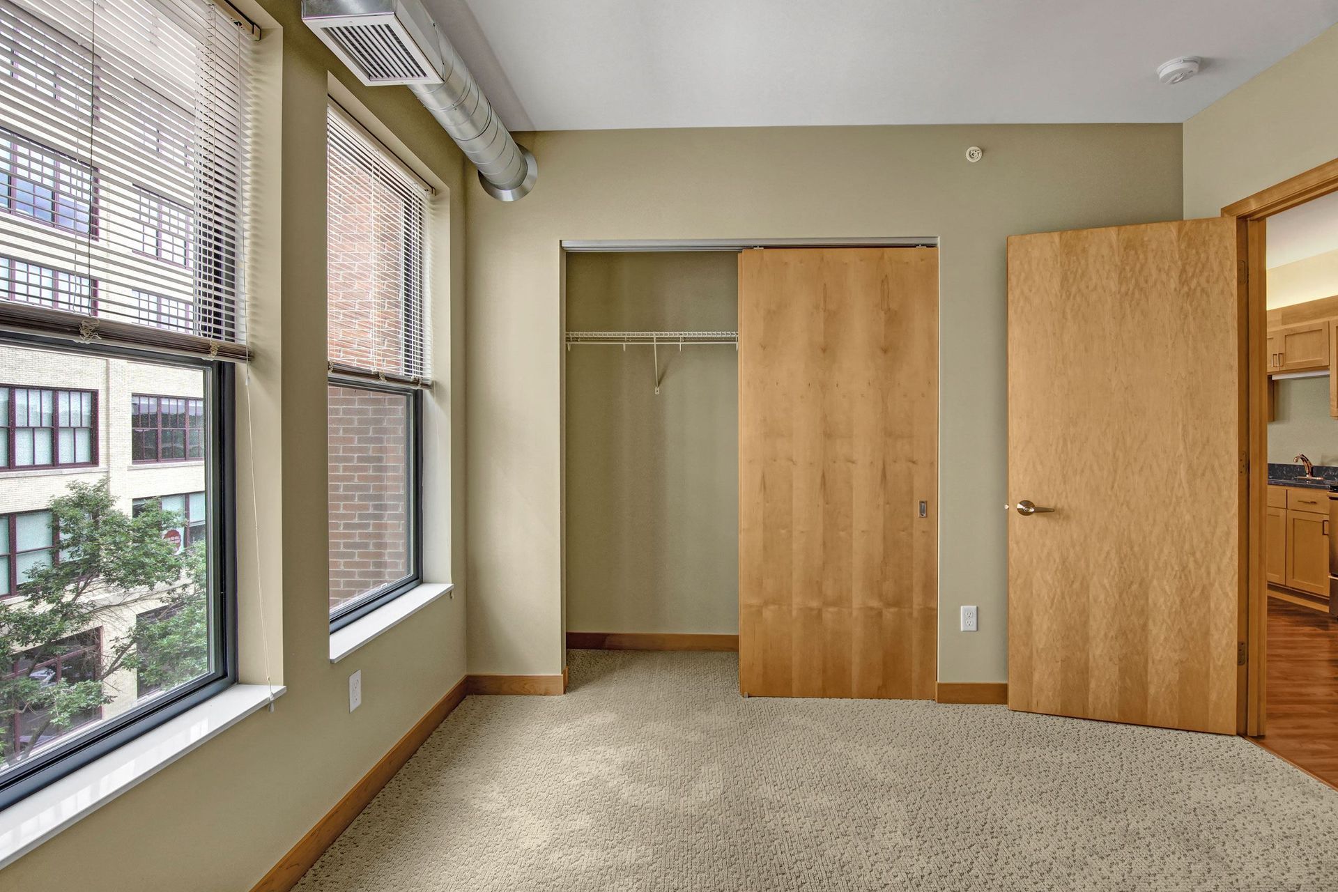Empty bedroom with windows, closet, and doorway to the right. Beige walls, light carpet, and light wood doors.