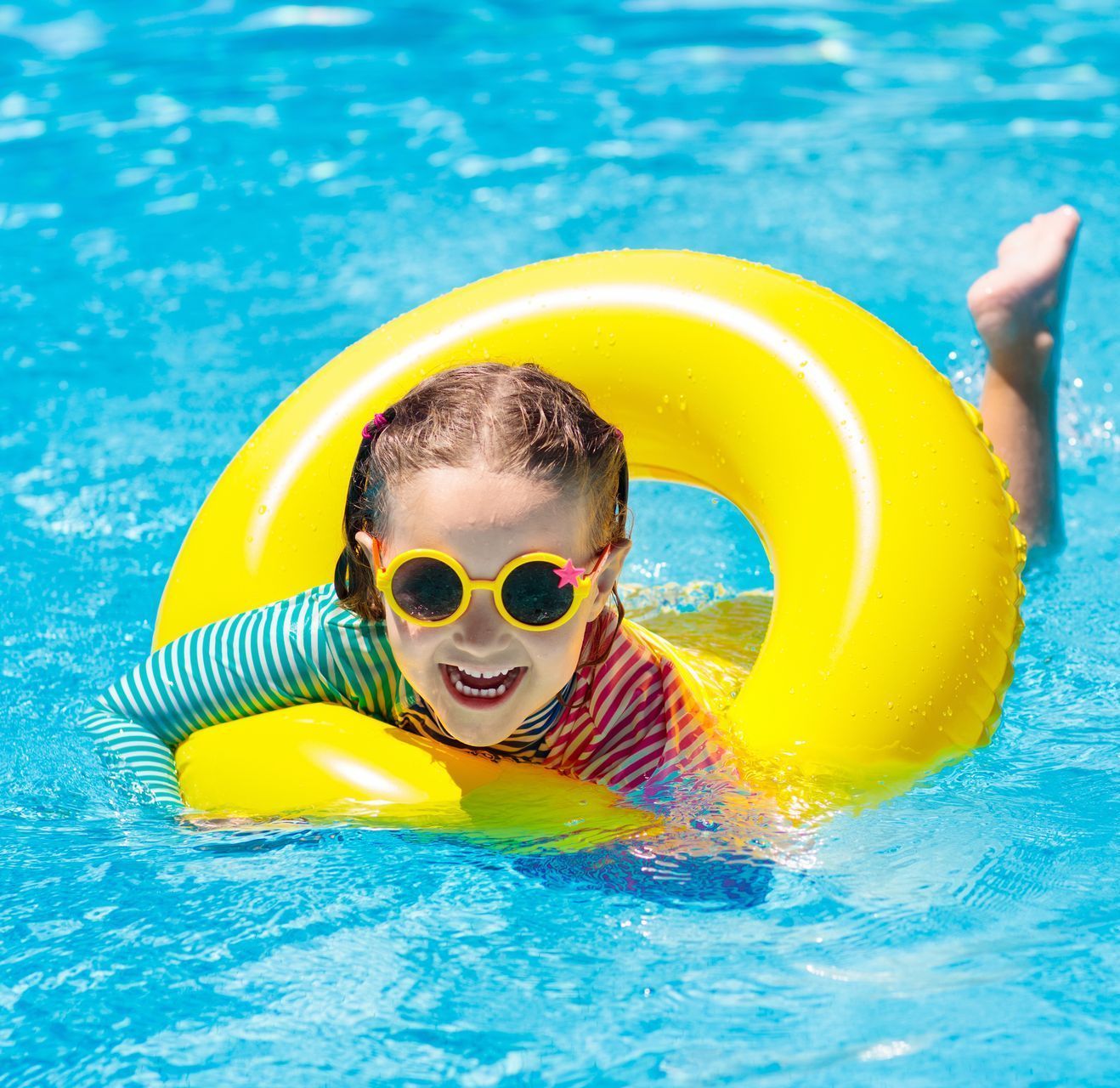 A young girl is swimming in a pool with a yellow floating ring around her. She is wearing sunglasses.