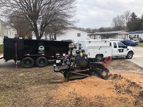 A dump truck is towing a stump grinder in a yard.