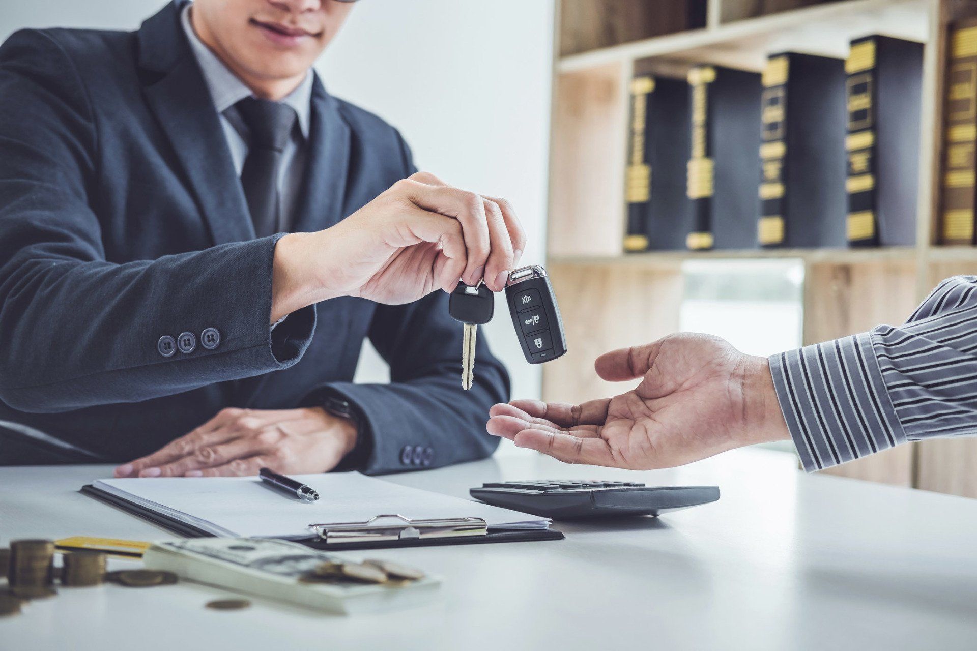 Man in suit hands car keys to another person at a desk, documents and calculator in front.