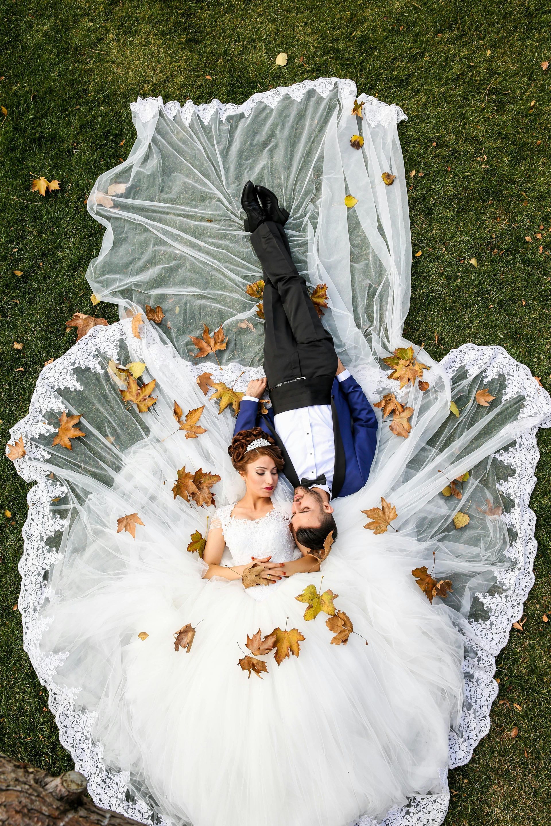 Bride and groom lying on grass, veil and dress spread, surrounded by autumn leaves.