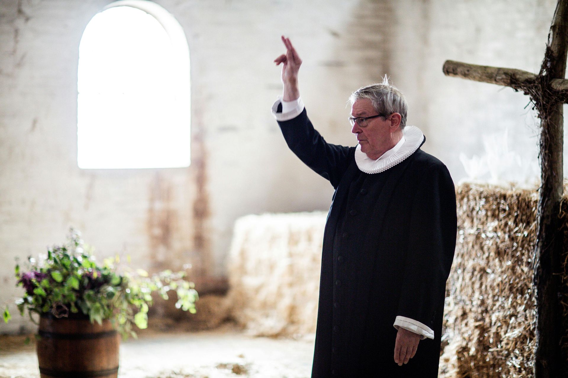 Clergyman in black robe raises hand near a window in a rustic interior, flowers in a barrel.