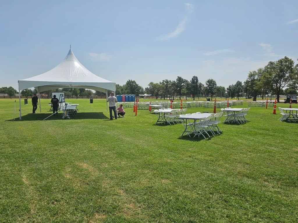 White tent, tables, on a grassy field under a blue sky. People are setting up.