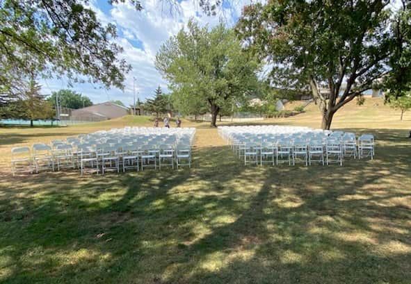 a row of white wedding chairs are lined up in a park .