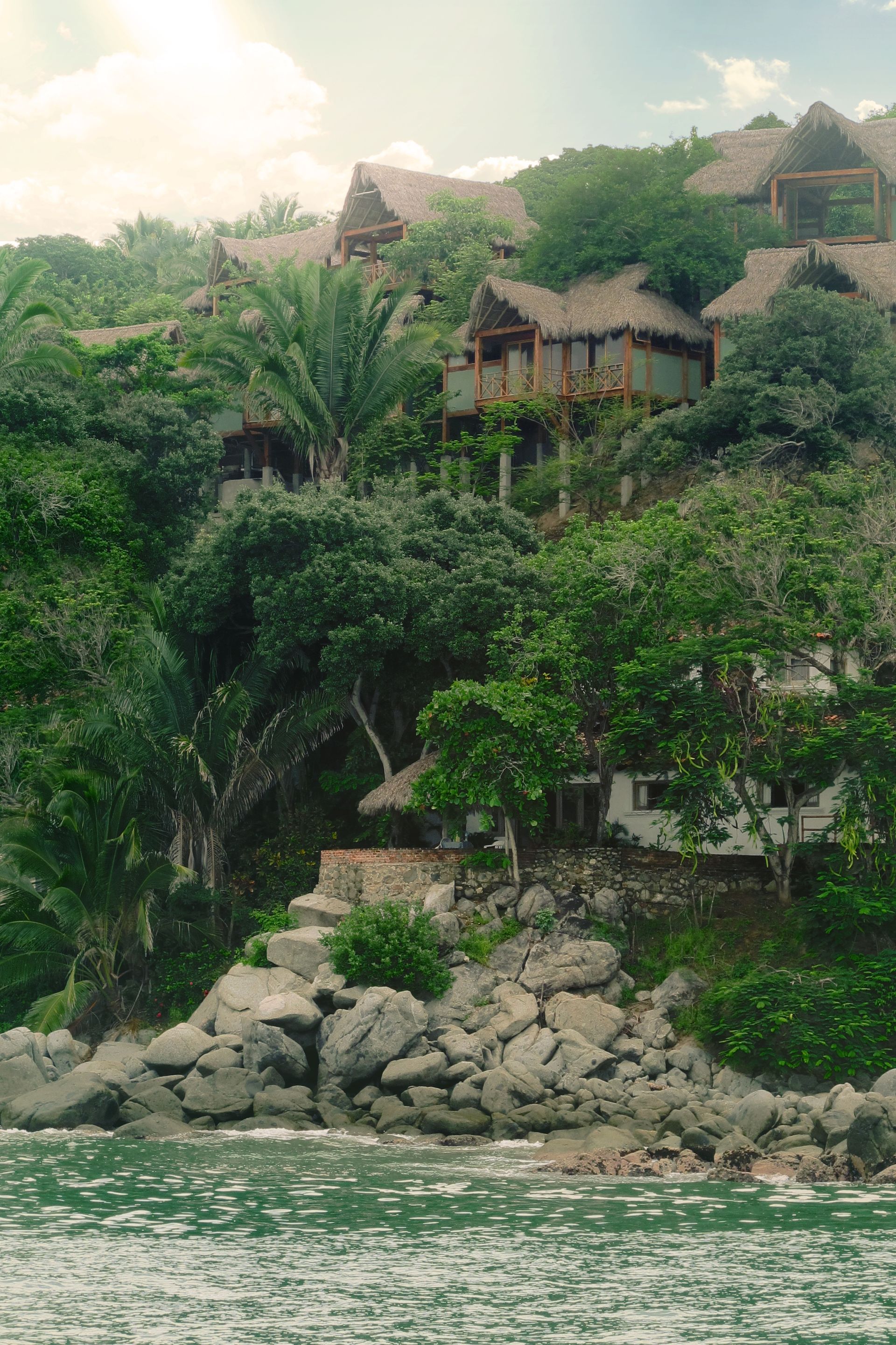 Cabañas con techo de paja en una exuberante ladera verde con vistas al agua.