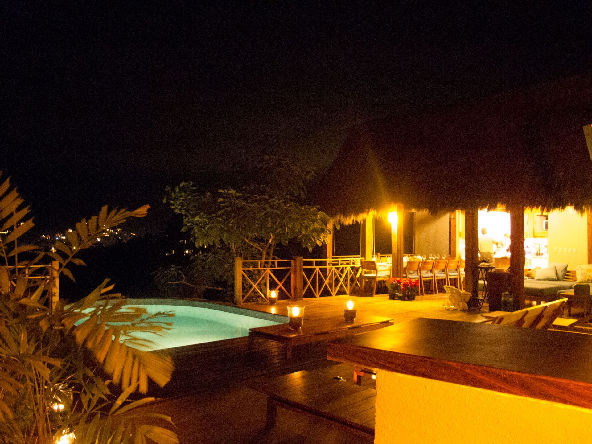 Vista nocturna de una piscina tropical y un patio con estructura de techo de paja, iluminado por luces cálidas.