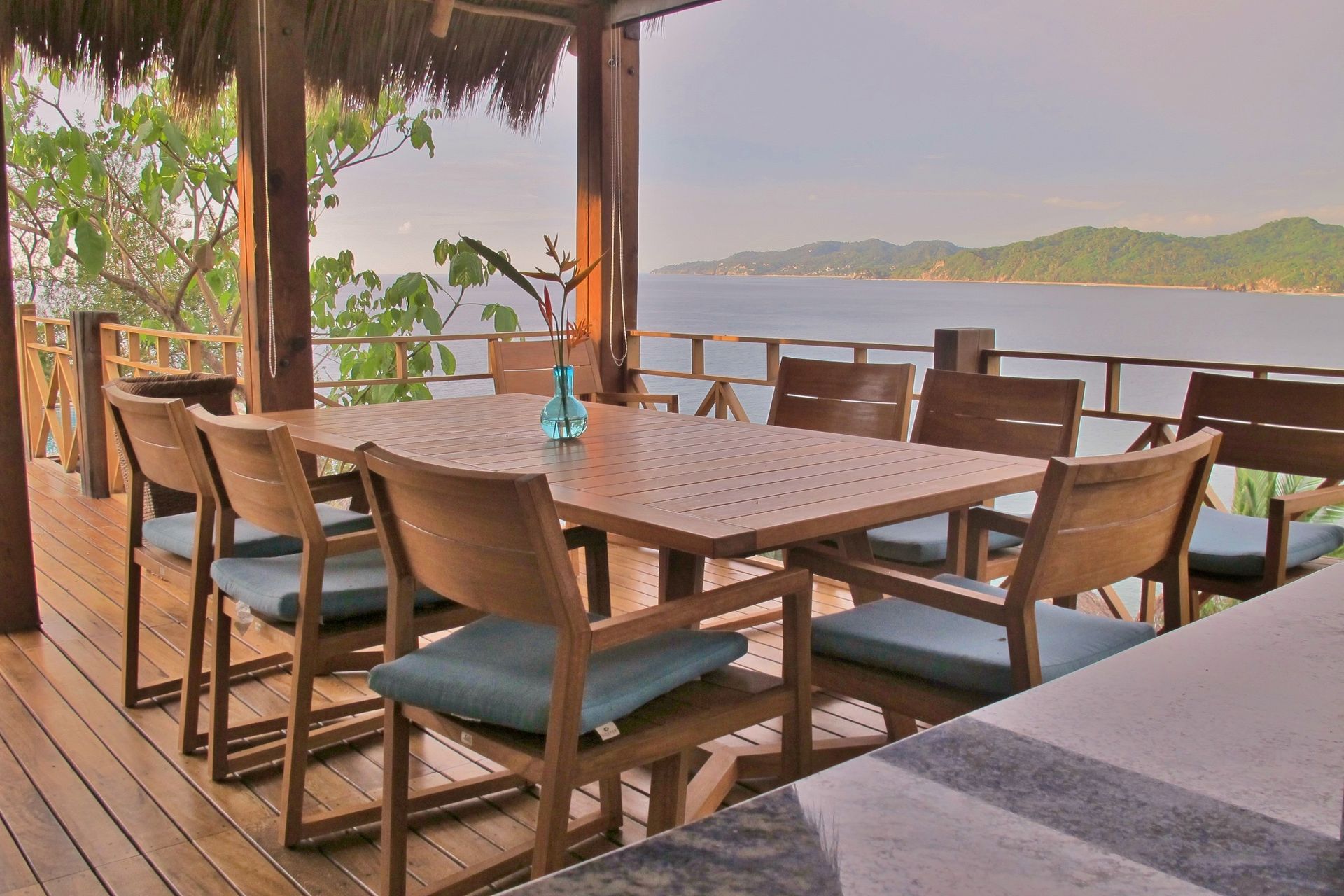 Mesa de comedor al aire libre con sillas en una terraza de madera con vista al océano.