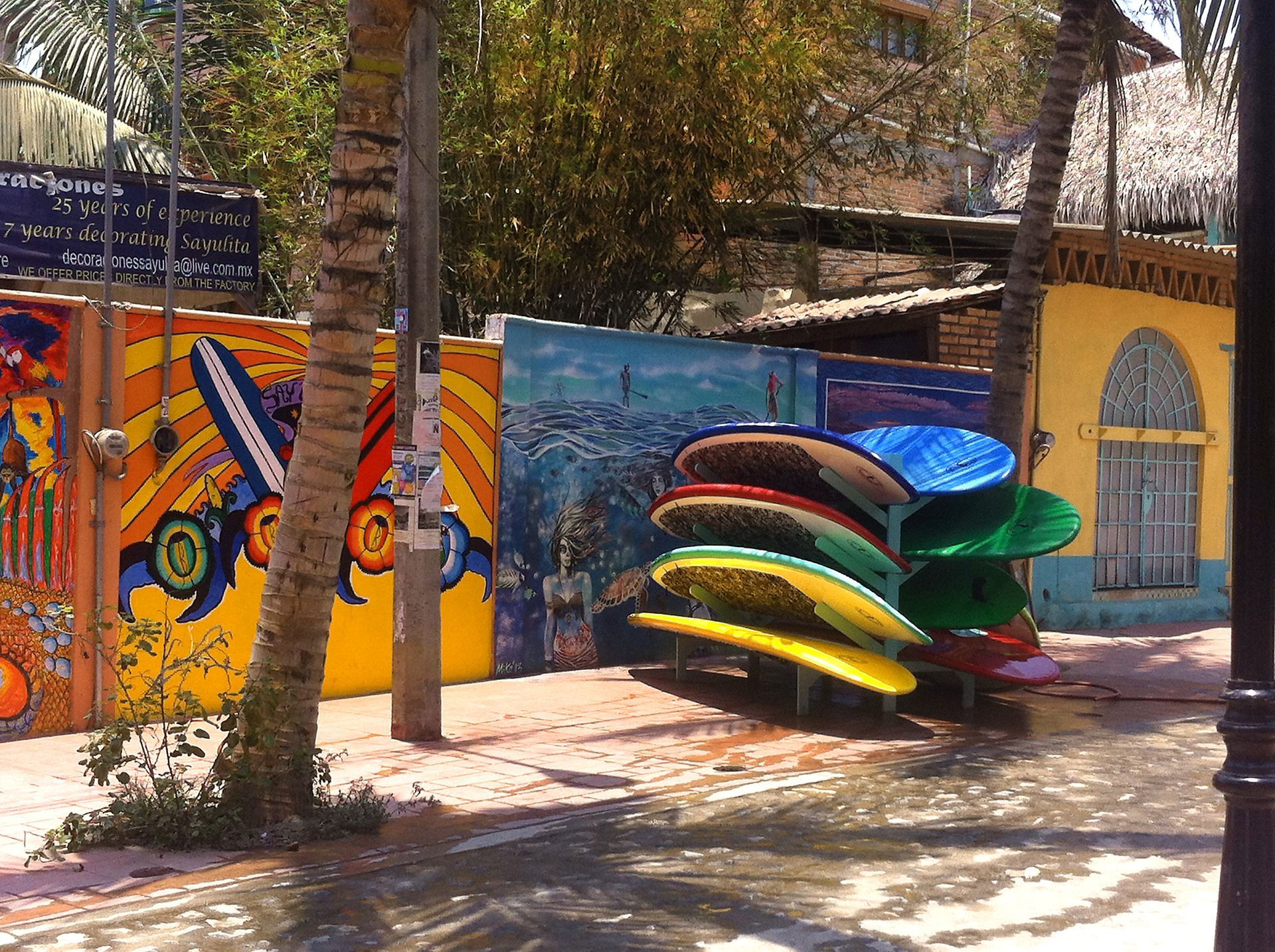 Tablas de surf coloridas apiladas junto a un mural con palmeras en un entorno tropical soleado.