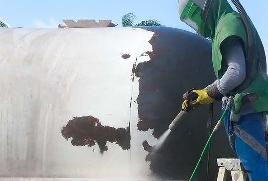 A man is sandblasting a large piece of metal.