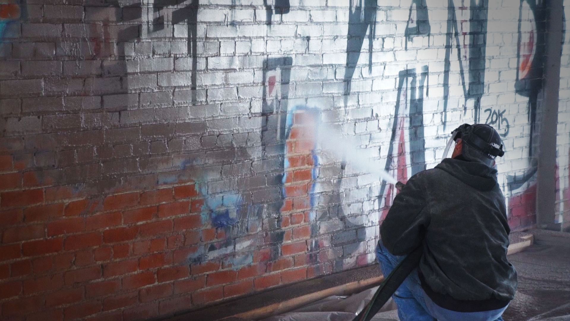 A man is cleaning graffiti on a brick wall with a high pressure washer.