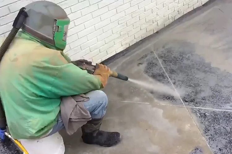 A man wearing a green shirt and a mask is using a high pressure washer on a concrete floor.