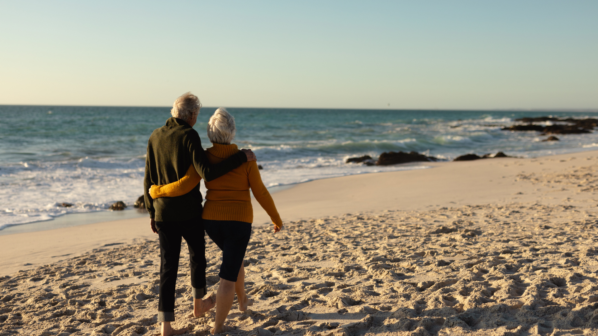 An elderly couple is walking on the beach looking at the ocean.