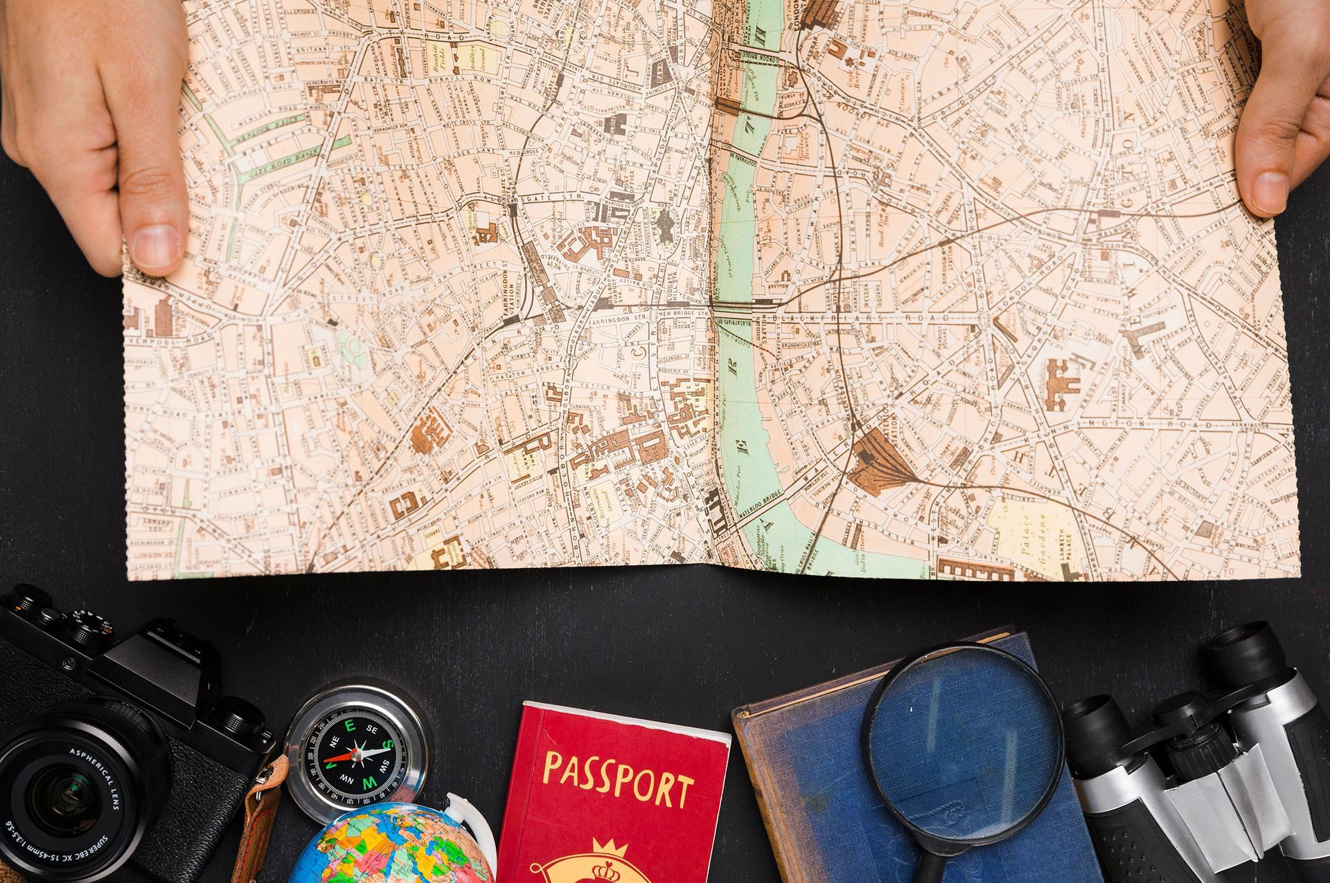 A person is holding a map next to a passport , compass , binoculars , and a camera.