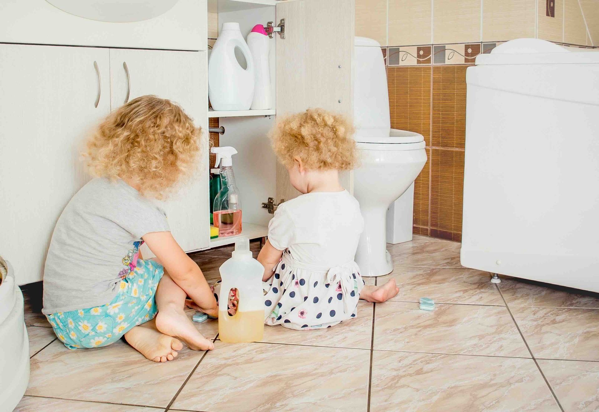 Two little girls are sitting on the floor in a bathroom next to a toilet.