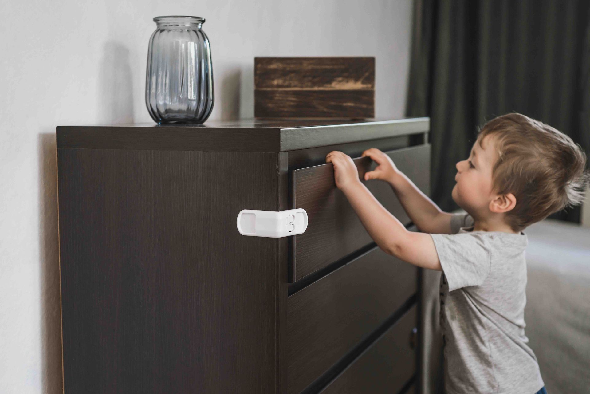 A young boy is playing with a dresser in a bedroom but the drawer is locked shut.