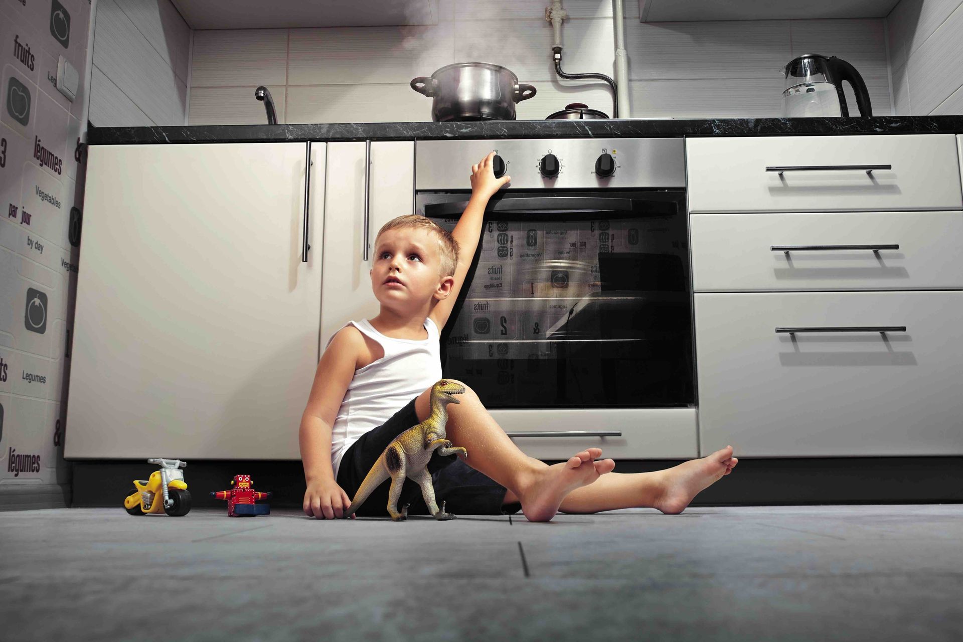 A little boy is sitting on the floor in a kitchen holding the oven knob and a toy dinosaur.