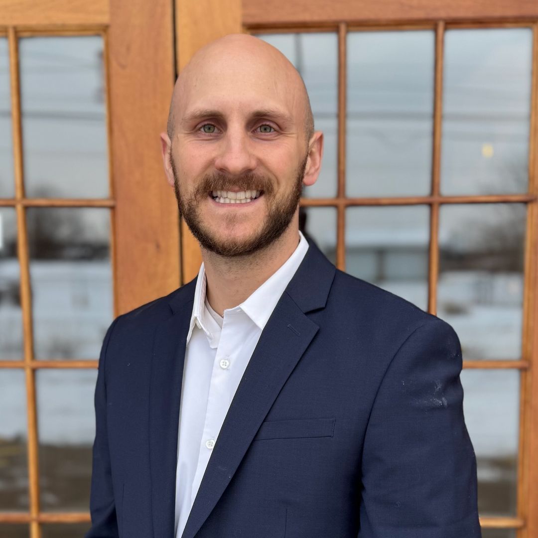 Cody Mahaffy in a suit and white shirt is smiling in front of a window.