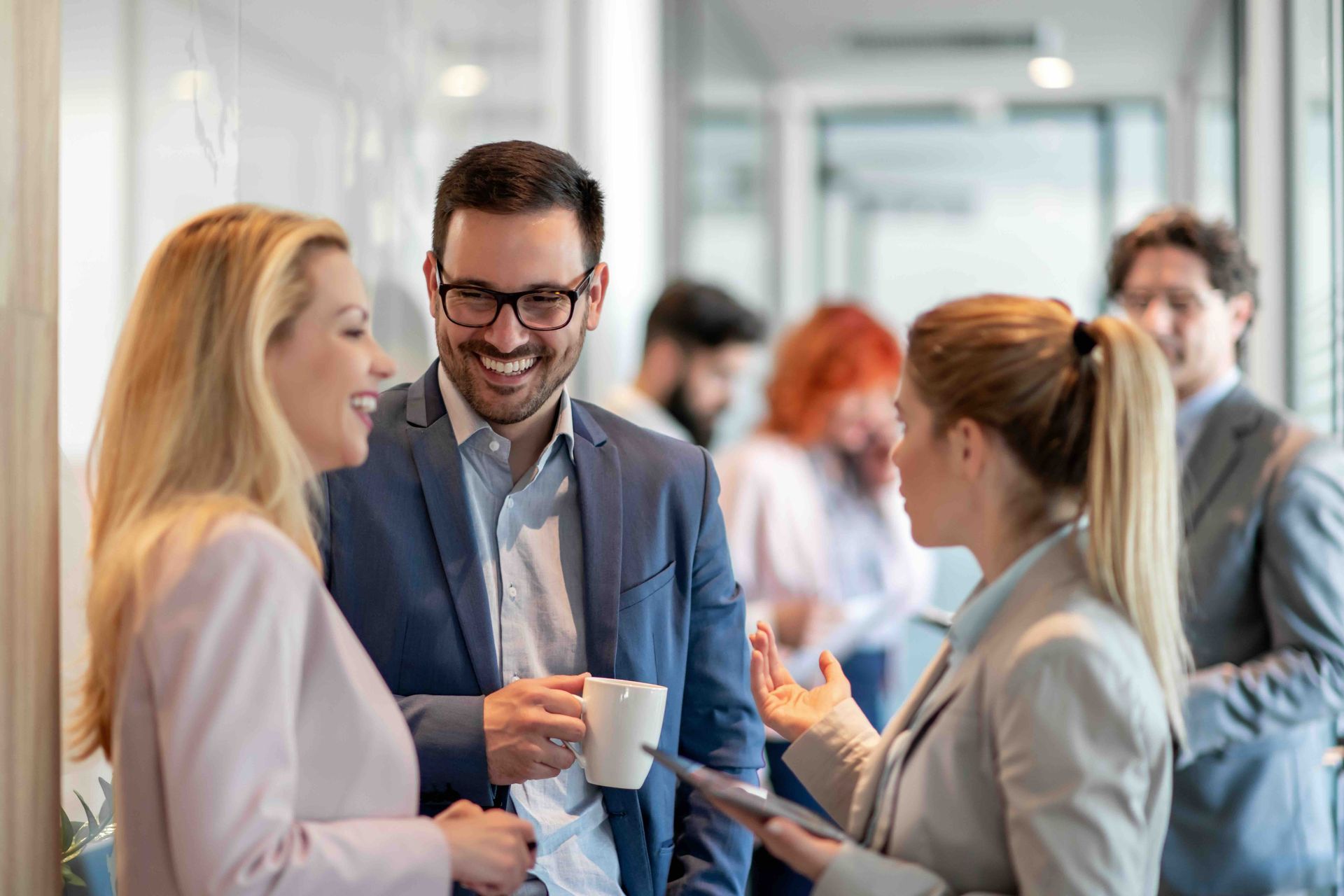 Office colleagues in suits chatting and smiling in a well-lit hallway. Man holding a mug.