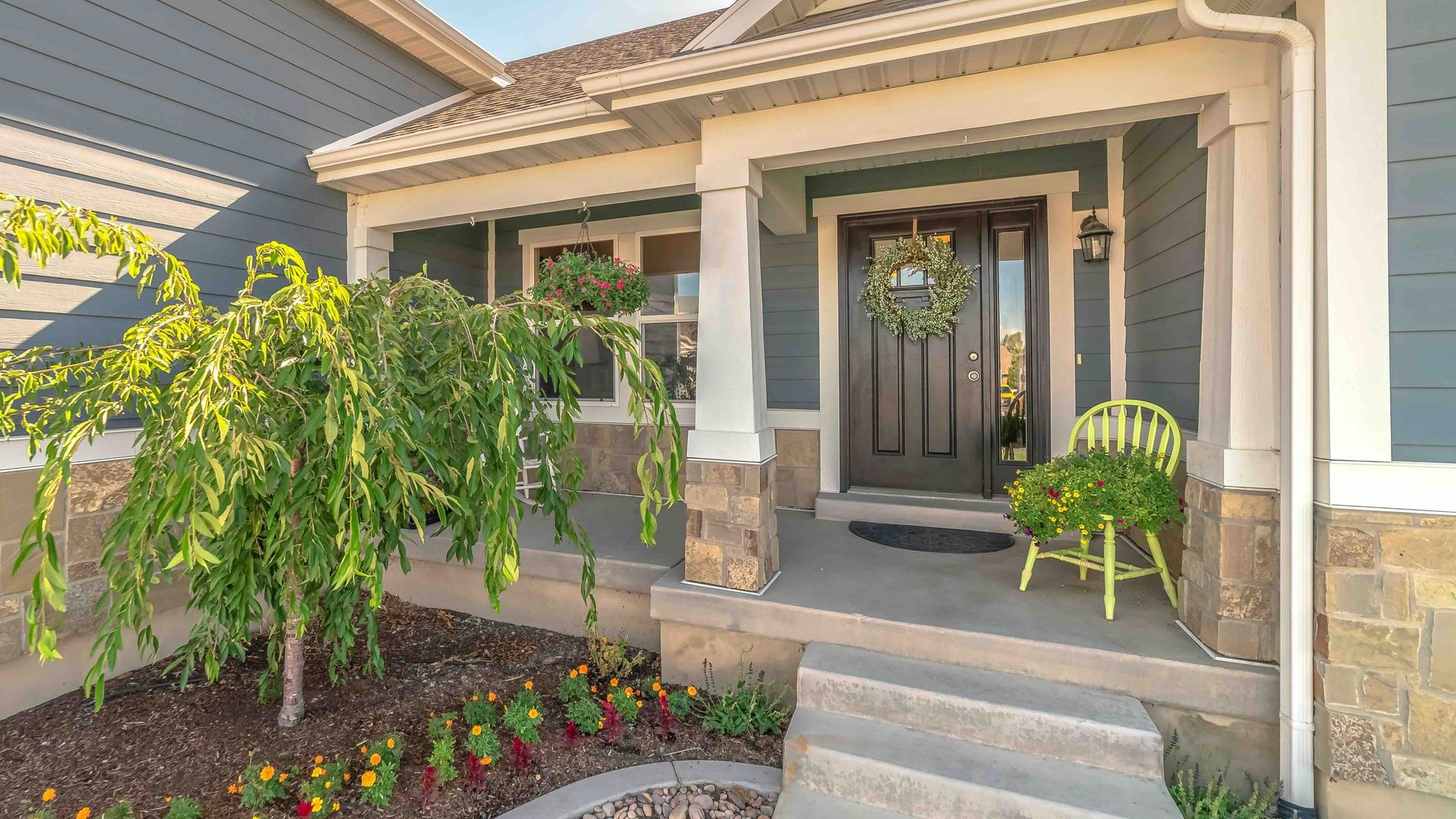 A blue-sided home with a stone-accented front porch, a wreath on the black door, a small tree, and a green chair.